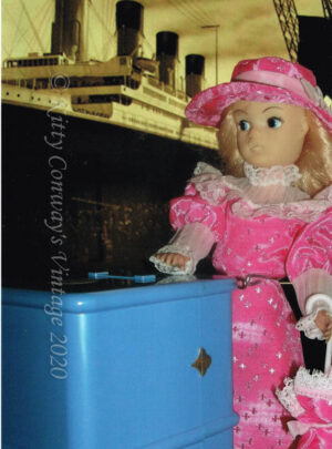 Blonde Sindy stands in a pink Edwardian style dress with her blue steamer trunk in front of an image of the Titanic